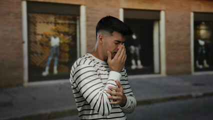 Fototapeta premium Young man with striped shirt standing on city street looking anxious in urban outdoor setting with hands near face expressing emotions under brick building backdrop.