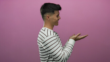 Young hispanic man smiling with hands extended to side against pink background in striped shirt conveying positive attitude, isolated for versatile use