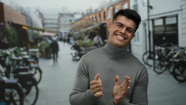 Young man clapping hands in a lively outdoor city street wearing glasses and a sweater with people and bicycles in the background showcasing a vibrant urban environment.