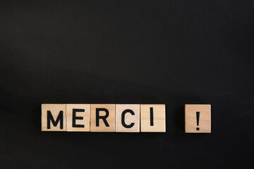 Wooden letter blocks spelling "MERCI!" on a black background, expressing gratitude in a minimalist and elegant composition. Thank you in French.