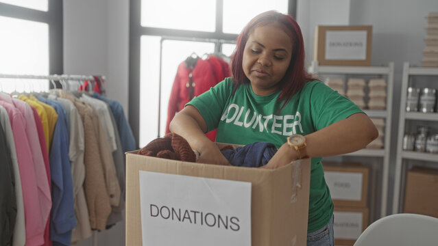 Woman volunteer folds navy sweater and places it into a cardboard donation box in building; generosity.