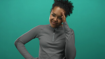 Fototapeta premium Dark skinned woman resting hand on temple with soft smile and tilting head slightly in turquoise studio; contemplation.