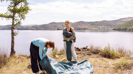 Two people set up a tent by a serene lake surrounded by hills in a calm setting © Sergey