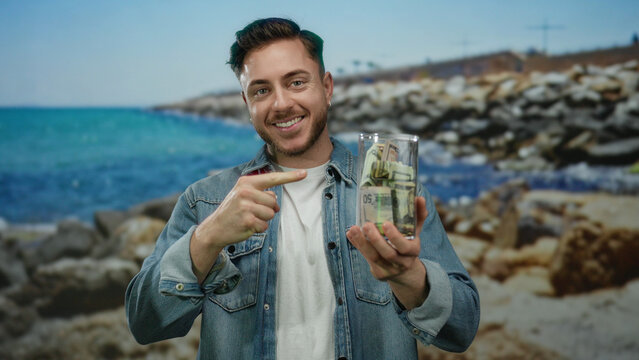 Young man at seaside holding money jar smiling with thumb up against scenic beach and ocean background expressing financial success outdoors