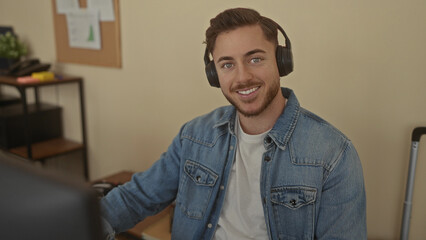 Young man with beard wearing headphones in an office setting, focused on work at computer in an indoor workplace environment, featuring casual attire and modern equipment.