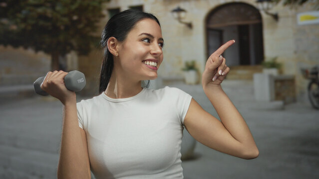 Hispanic woman exercising with dumbbell outdoors smiling in sunny urban area pointing aside confidently - Powered by Adobe