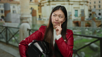 Fototapeta premium Woman in red jacket holds helmet while pondering in front of ancient roman ruins in city setting, combining modern and historical elements outdoors.