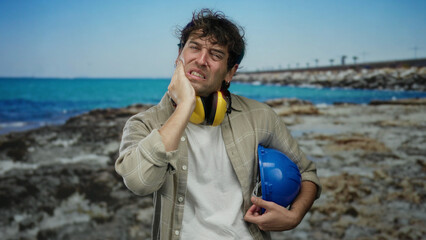 Hispanic man wearing headphones and hardhat stands on a beach holding his cheek in discomfort, with...