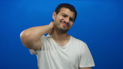 Young hispanic man holding neck with pained expression against vivid blue wall in studio;...