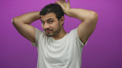 Young hispanic man making antler ear gesture with hands on head in studio with purple backdrop; playfulness fun joy whimsy.