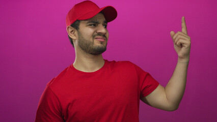 Young hispanic delivery man in red shirt and cap gestures thumb down with his finger in magenta...