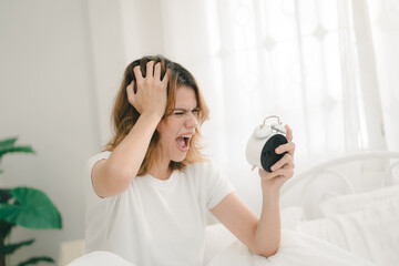 Frustrated woman screaming at alarm clock in bed, showing stress from waking up late or sleep issues, perfect for concepts of anxiety, oversleeping, morning stress, or burnout.