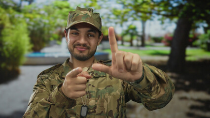 Man soldier in camouflage military uniform makes l hand gesture toward camera in forest park;...
