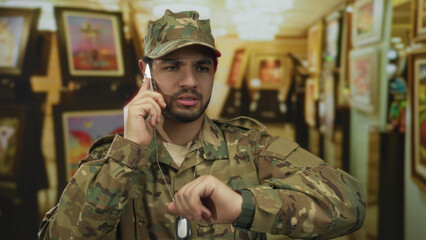 Man in camouflage uniform holding phone to ear checks wristwatch in building lined with paintings;...