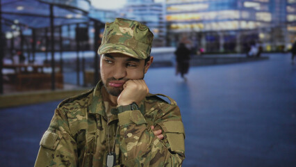 Fototapeta premium Hispanic soldier in camouflage uniform with dog tags stands with arms crossed on a city street; doubt.