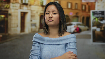 Fototapeta premium Young chinese woman with serious expression standing on a city street, wearing a blue sweater and looking away, with blurred urban background.