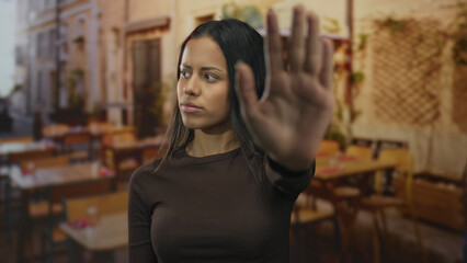 Woman gesturing stop on a restaurant terrace, street background, suggested outdoors, young hispanic female showing hand, casual setting, blurred chairs visible.