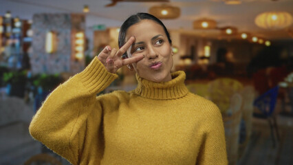 Fototapeta premium Young hispanic woman in yellow sweater makes playful peace sign inside a warmly lit hotel indoors, showcasing a joyful and friendly atmosphere.