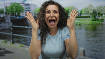 Young woman in blue shirt covers eyes with hands on street beside canal waters; surprise curiosity...