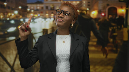 Young african american woman wearing glasses with subtle smile and arms crossed on city street at night; rejection.