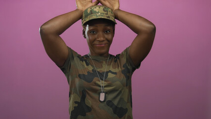 Woman soldier in camouflage cap and dog tags raises hands in pink studio; playful confidence energy...