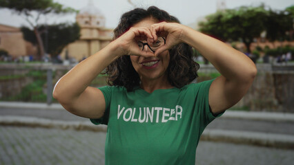 Woman wearing green volunteer shirt making heart shape with hands in front of scenic roman ruins...