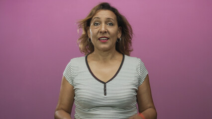 Hispanic middleage woman extending arms and pointing both fingers toward camera in pink studio; warm invitation embrace.