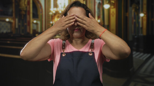 Fototapeta Middle age hispanic woman in pink shirt and apron covers eyes with hands in church building  serenity.