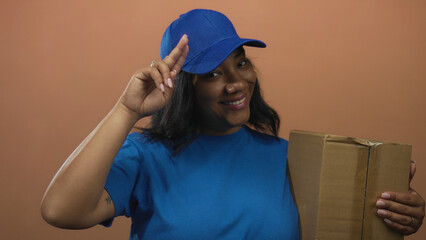 Delivery woman holds cardboard box and salutes head with fingers against peach wall in studio scene; pride duty.