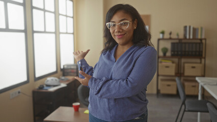 African american woman gestures with both hands toward large window in office building; confidence empowerment.