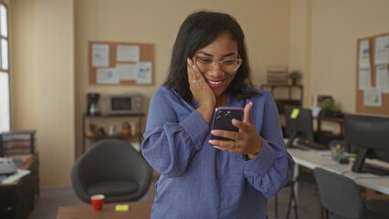 African american woman smiling while tapping smartphone at wooden desk inside office building with computer monitors and cup; personal joy.