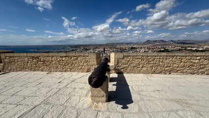 Cannon on the Horizon: A weathered cannon, positioned on a stone terrace, surveys a panoramic vista of a coastal town under a vast, vibrant sky, the scene rich with history and grandeur.