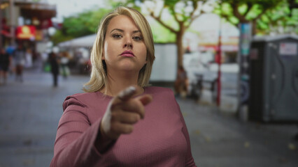 Fototapeta premium Woman with blonde hair holds out her index finger and points forward on a crowded urban street under daylight; assertiveness.