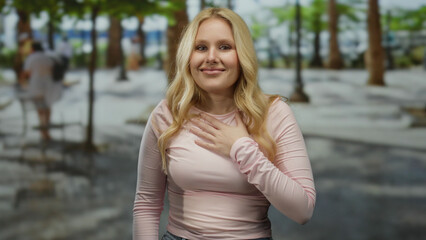 Obraz premium Young blonde woman on a city street outdoors expressing gratitude with hand on chest under green trees creating a serene urban atmosphere of happiness and appreciation