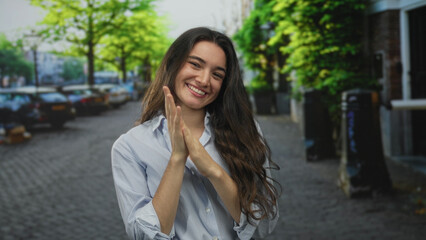 Fototapeta premium Woman claps hands together in front of chest on sunlit street with parked cars and trees; happiness gratitude.