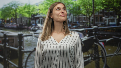 Fototapeta premium Woman stands on street by canal under green trees and parked bicycle near brick buildings; serenity.