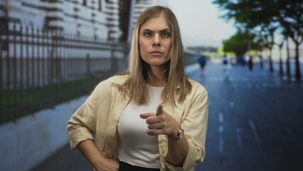 Determined young blonde woman points index finger toward camera on sunny urban street; confidence.