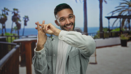 Young hispanic man making hashtag gesture on sunny seaside promenade with ocean view and palm trees, exuding happiness and relaxation outdoors.