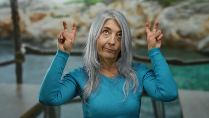 Woman expressing joy at seaside outdoor with peace gestures, grey hair gently flowing in beach breeze, capturing serene and relaxed senior vibe in natural setting.