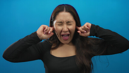 Young hispanic woman with long dark hair covers ears with hands on a vibrant blue studio set;...