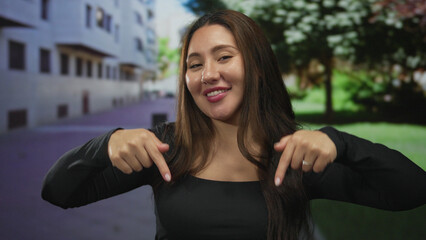 Fototapeta premium Hispanic woman wearing black top points both fingers downward on street amid green trees; invitation.