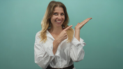 Woman points finger to palm in studio teal backdrop wearing white shirt and belt; cheerful confidence.