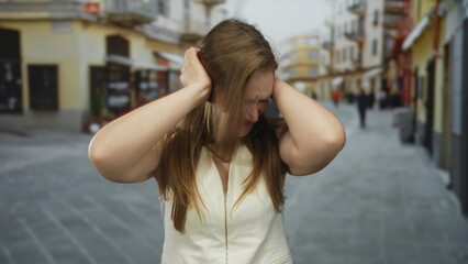 Fototapeta premium Caucasian woman covering her ears with both hands while grimacing on a busy street lined with buildings; frustration.