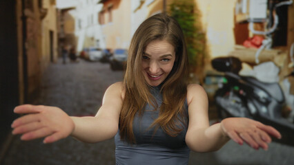 Young caucasian woman extends hands in a cheerful come here gesture on a cobblestone street; warm friendly welcome invitation.