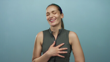 Woman laughing with hand pressed to chest wearing sleeveless top against a light blue studio wall;...
