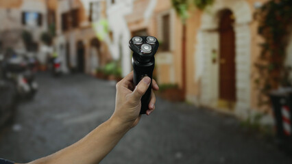 Man holding electric shaver on a cobblestone street in a rustic town, showcasing grooming in an...