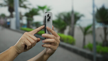 Man using smartphone outdoors in a city street, showcasing modern technology and digital communication in an urban environment with blurred background trees.
