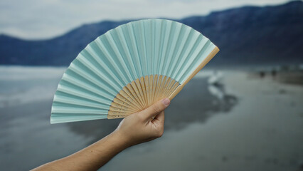 Caucasian man holding a light blue hand fan on a serene seaside beach with mountains in the background, showcasing a peaceful outdoor setting.