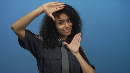 Woman in police uniform with grey tie framing smiling face with hands against blue wall in studio;...