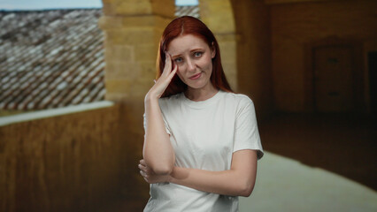 Woman standing in outdoor university courtyard with hand on head, appearing thoughtful and reflective, showcasing a serene and picturesque academic environment.